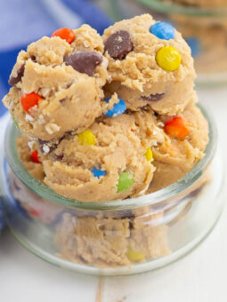 Scoops of cookie dough with colorful candy-coated chocolates and chocolate chips in a small glass jar, next to a blue and white checkered cloth.