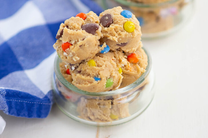 Scoops of cookie dough with colorful candy-coated chocolates and chocolate chips in a small glass jar, next to a blue and white checkered cloth.