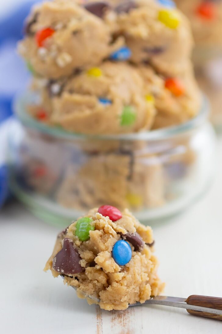 A close-up of a spoonful of cookie dough with chocolate chunks and colorful candy pieces, with more cookie dough in a glass jar in the background.