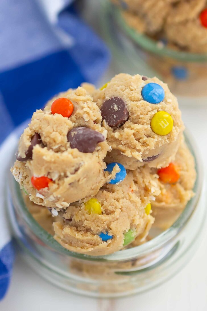 Scoops of cookie dough with colorful chocolate candies and chocolate chips in a glass jar, with a blue and white cloth in the background.