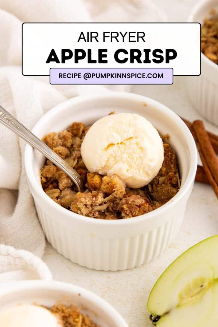 A small white ramekin holds apple crisp topped with a scoop of vanilla ice cream and a spoon. Sliced green apple and cinnamon sticks rest nearby. Text reads: Air Fryer Apple Crisp. Recipe @pumpkinnspice.com..