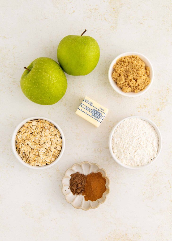Two green apples, a pat of butter, a small bowl of brown sugar, a bowl of oats, a bowl of flour, and a dish with cinnamon and nutmeg are arranged on a light-colored surface.