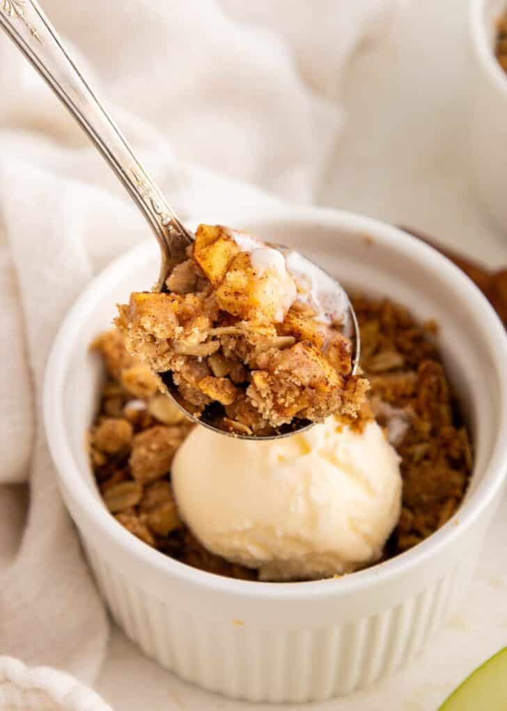A spoonful of apple crisp hovers over a white ramekin filled with more apple crisp and a scoop of vanilla ice cream.