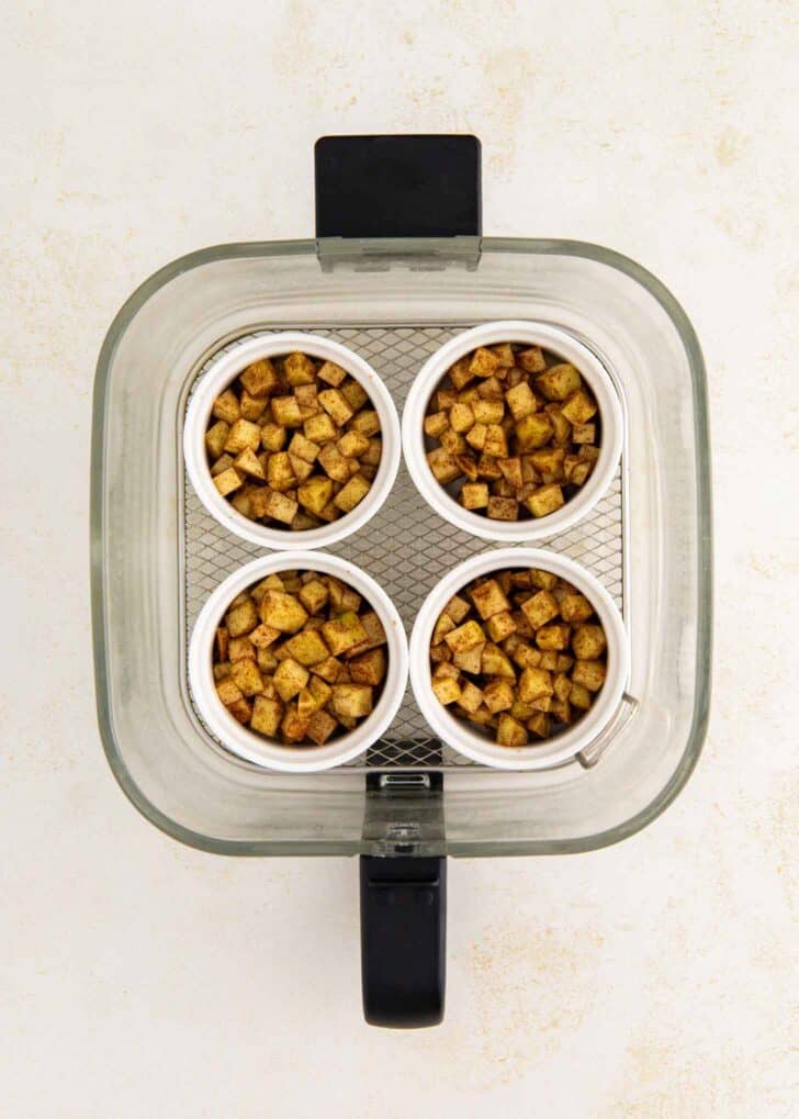 Top view of four small white ramekins filled with spiced apple cubes, arranged in a glass air fryer basket on a light-colored surface.