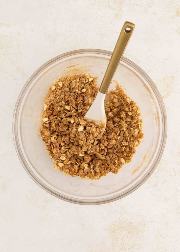 A glass bowl with crumbly oat mixture and a spoon, viewed from above, on a light surface.