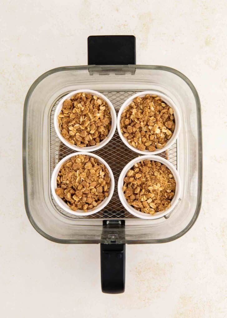 Four small ramekins filled with crumbly oat topping and apples are arranged in a square inside an air fryer basket, viewed from above on a light background.