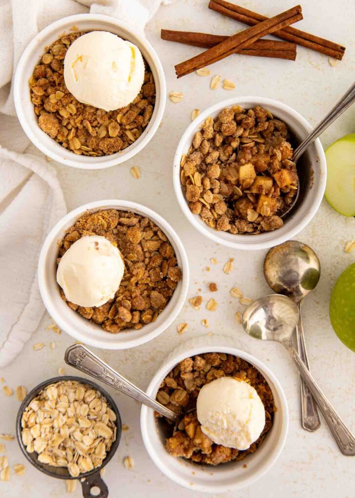 Four ramekins of apple crisp topped with vanilla ice cream, surrounded by oats, green apple slices, cinnamon sticks, and three spoons on a white surface.