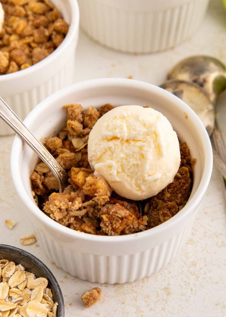 A white ramekin filled with apple crisp, topped with a scoop of vanilla ice cream and a spoon. Crumbled oats and another ramekin are visible nearby.