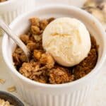 A white ramekin filled with apple crisp topped with a scoop of vanilla ice cream. A spoon rests in the dish, and there are apples, oats, and spoons nearby.