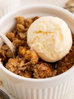 A white ramekin filled with apple crisp topped with a scoop of vanilla ice cream. A spoon rests in the dish, and there are apples, oats, and spoons nearby.