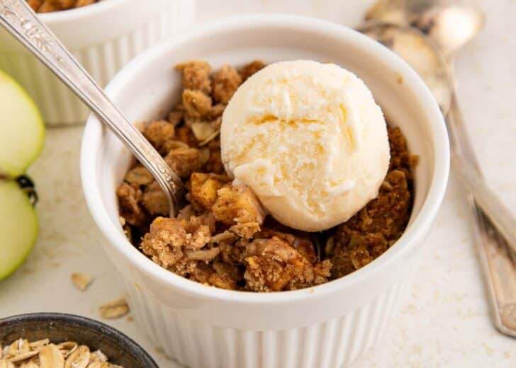 A white ramekin filled with apple crisp topped with a scoop of vanilla ice cream. A spoon rests in the dish, and there are apples, oats, and spoons nearby.