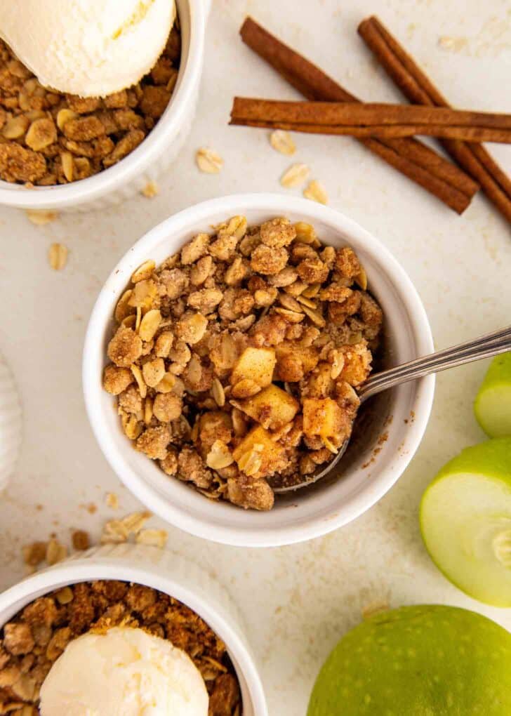 Mini apple crisps with oat topping in ramekins, one with a spoon inside, served with vanilla ice cream. Green apple slices and cinnamon sticks are nearby on a white surface.