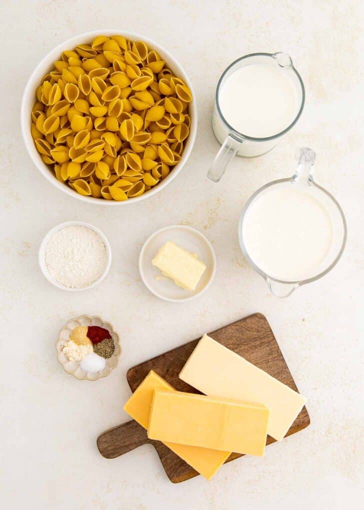Dry pasta shells, two measuring cups of milk, a small bowl of flour, a pat of butter, spices in a small dish, and several blocks of cheese on a wooden board are arranged on a white surface.