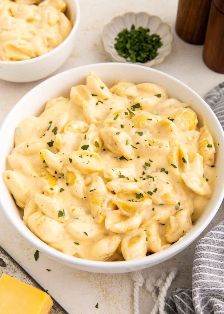 A white bowl filled with creamy macaroni and cheese shells, garnished with chopped herbs. A small dish of herbs, a striped napkin, and blocks of cheese are nearby.