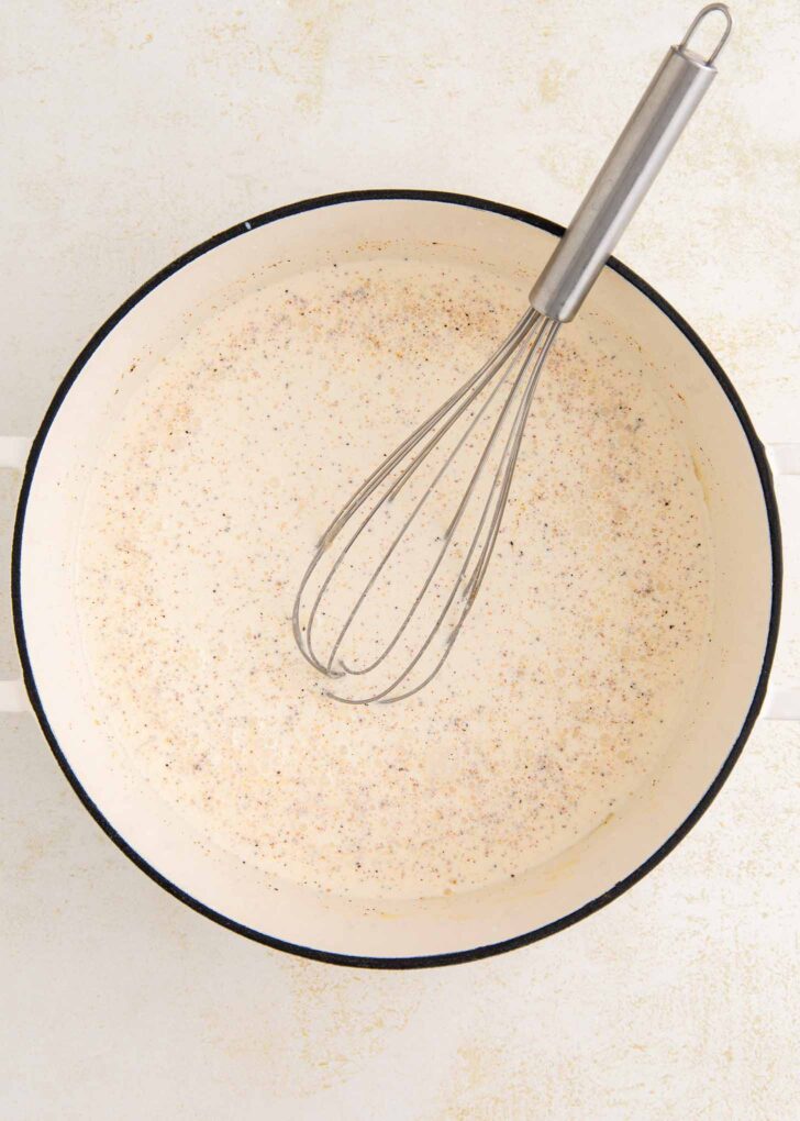 A metal whisk rests in a white, round pot containing a creamy liquid speckled with black pepper, placed on a light-colored surface.