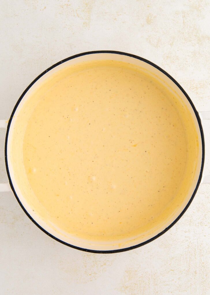 A white pot filled with creamy, light orange cheese sauce sits on a light-colored surface.