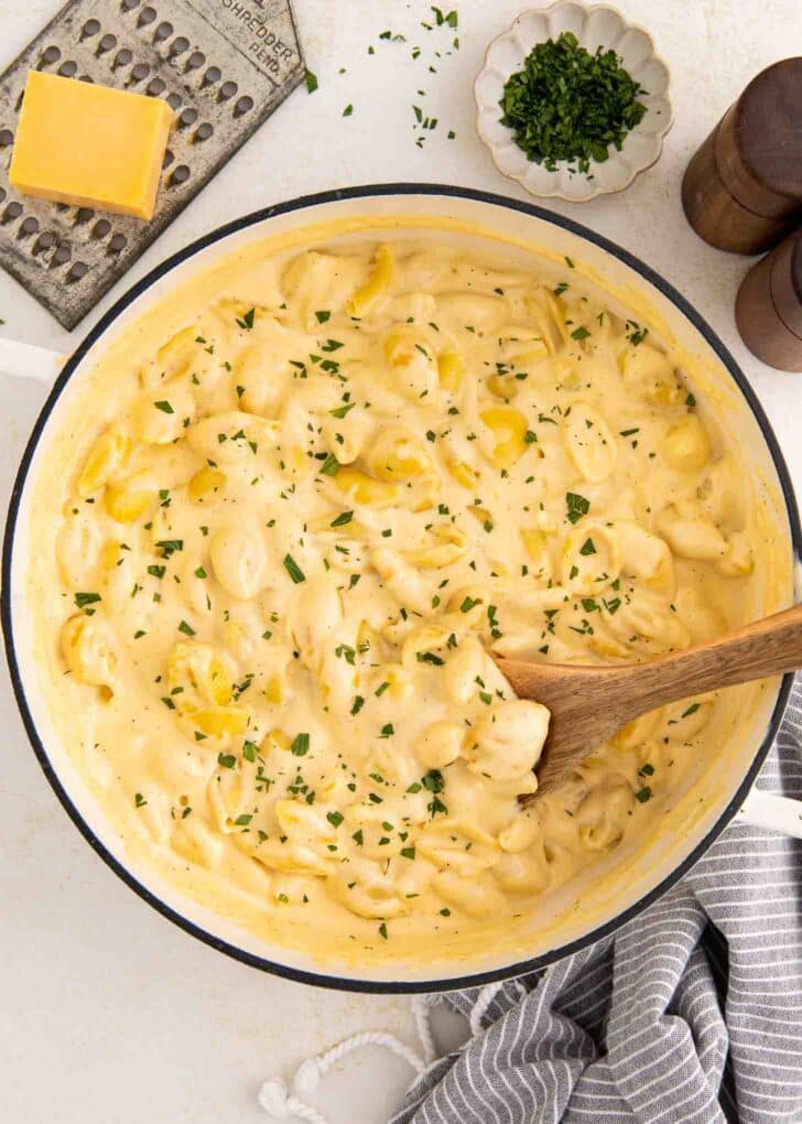 Creamy cheese elbow noodles in a large white pot, garnished with chopped herbs. A wooden spoon rests in the pot. Nearby are a cheese grater, a block of cheese, a dish of herbs, salt and pepper shakers, and a striped towel.