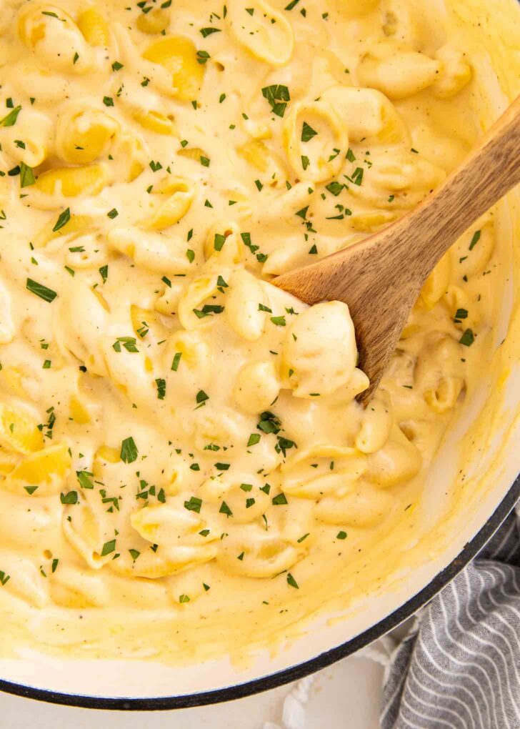 Creamy macaroni and cheese with shell pasta in a pot, topped with chopped herbs, being stirred with a wooden spoon.
