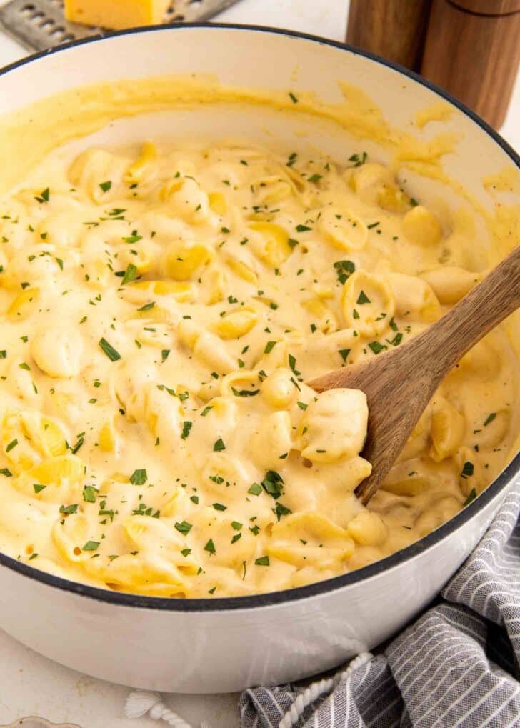 A pot filled with creamy macaroni and cheese topped with chopped herbs. A wooden spoon is stirring the pasta, and a gray-striped towel rests beside the pot.