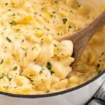 Creamy macaroni and cheese with shell pasta in a pot, topped with chopped herbs. A wooden spoon is stirring the cheesy mixture. A striped towel is partially visible beside the pot.