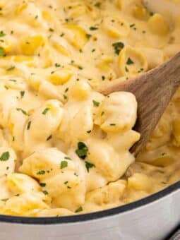 Creamy macaroni and cheese with shell pasta in a pot, topped with chopped herbs. A wooden spoon is stirring the cheesy mixture. A striped towel is partially visible beside the pot.