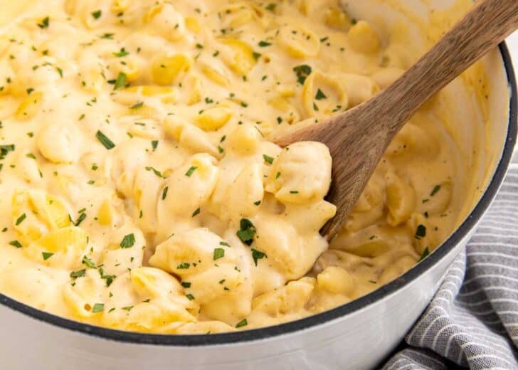Creamy macaroni and cheese with shell pasta in a pot, topped with chopped herbs. A wooden spoon is stirring the cheesy mixture. A striped towel is partially visible beside the pot.