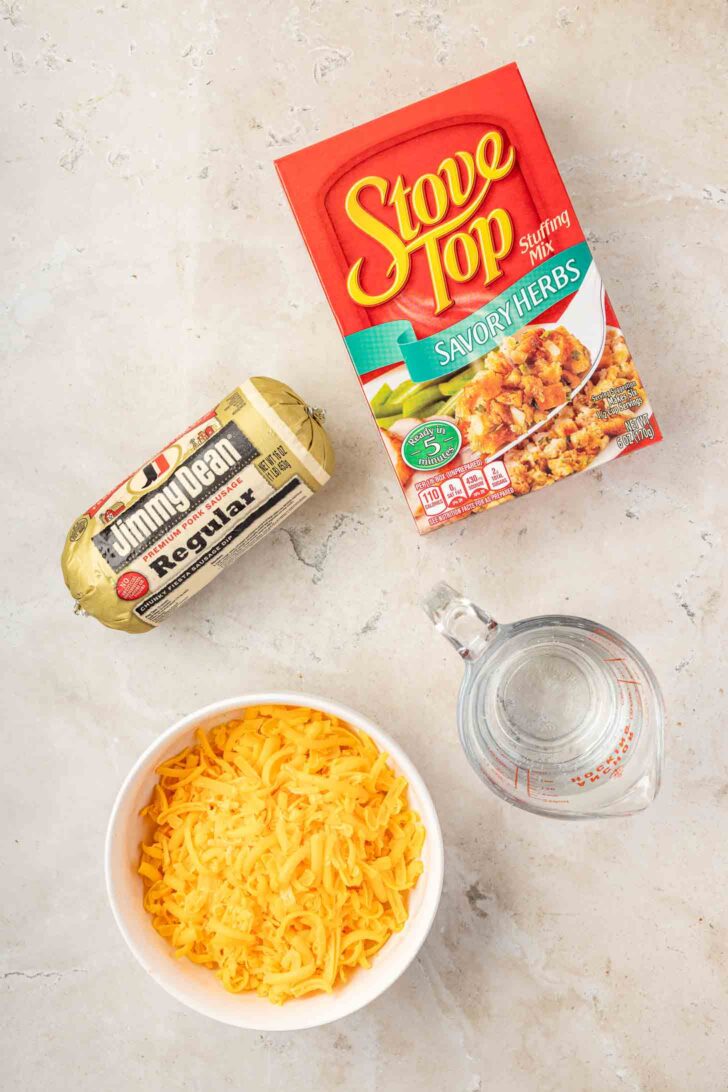 Ingredients on a countertop: a package of Stove Top Savory Herbs stuffing mix, a tube of Jimmy Dean regular sausage, a bowl of shredded cheddar cheese, and a glass measuring cup with water.