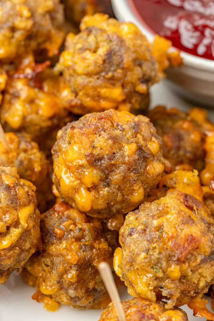 Several golden-brown stuffing balls with melted cheese are stacked closely together, with a bowl of red dipping sauce visible in the background. Some sausage balls have wooden toothpicks inserted.