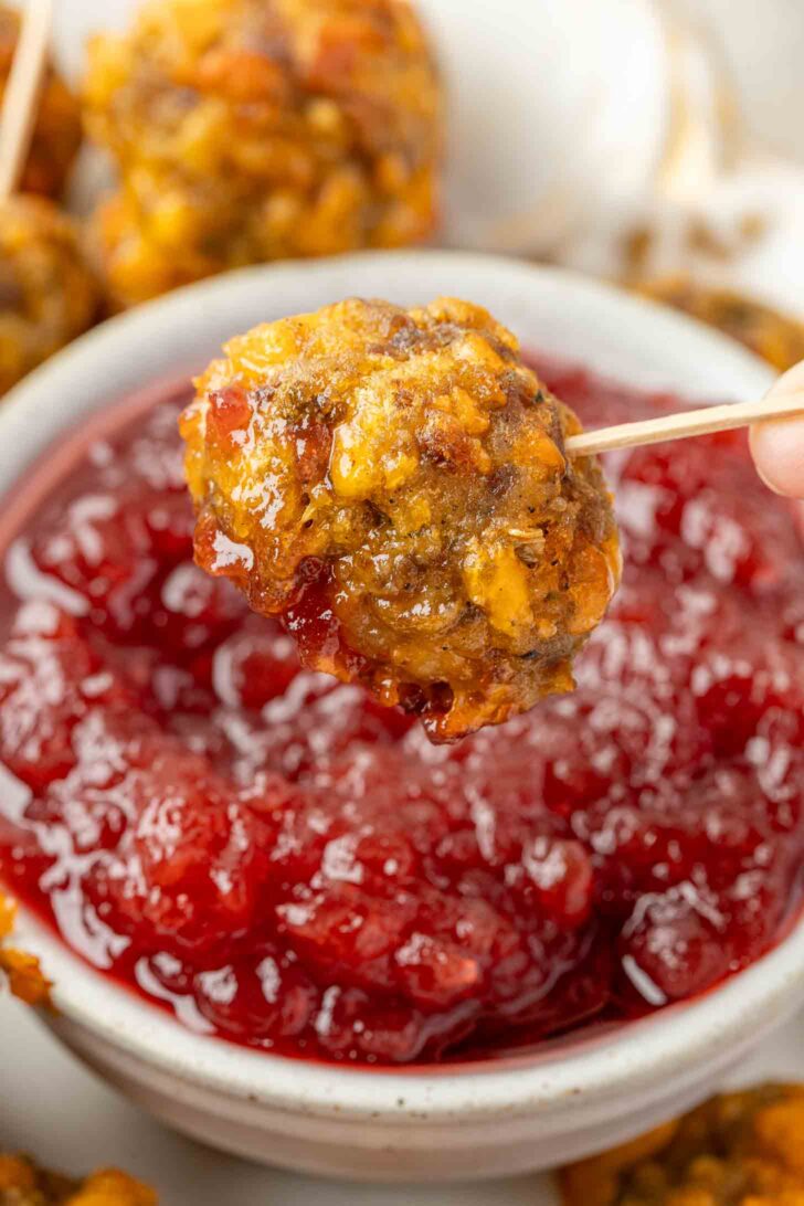 A hand holds a toothpick with a stuffing balls dipped in glossy red cranberry sauce, with more sauce and meatballs visible in the background.