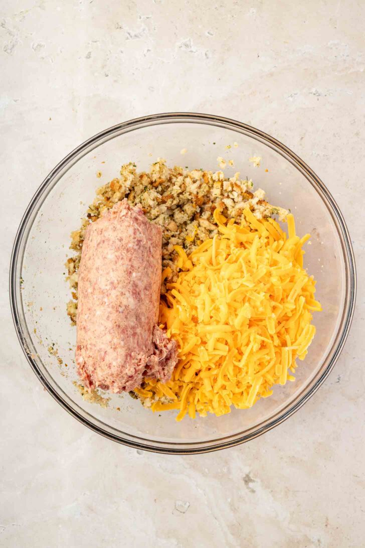 A clear bowl contains ground meat, shredded cheddar cheese, and stuffing mix, all unmixed, on a light-colored surface.