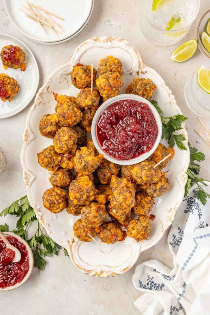 A platter of stuffing balls surrounds a bowl of cranberry sauce, garnished with parsley. Toothpicks are nearby, with a few stuffing balls and extra sauce on side plates.