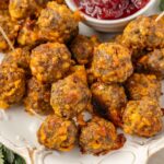 A plate of baked stuffing balls, some with toothpicks, sits next to a small bowl of red cranberry. Fresh herbs decorate the edges of the plate.