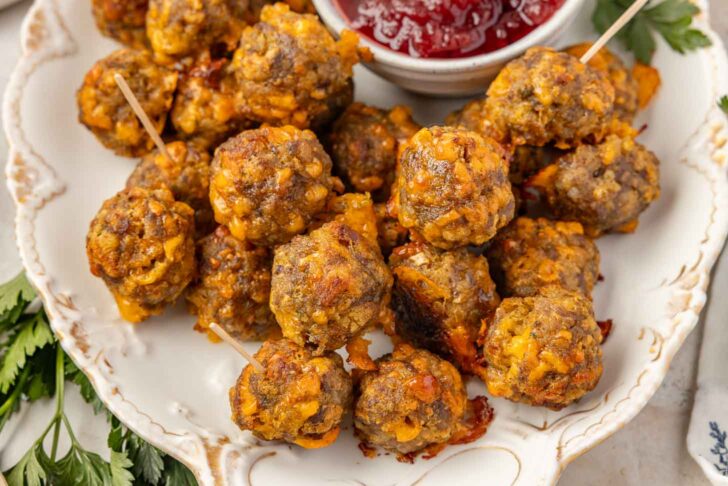 A plate of baked stuffing balls, some with toothpicks, sits next to a small bowl of red cranberry. Fresh herbs decorate the edges of the plate.