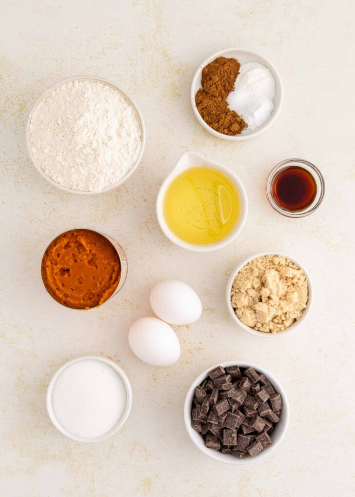 Bowl of flour, bowl spices, baking soda and salt, oil, vanilla, brown sugar, white sugar, pumpkin puree, two eggs, and chocolate chunks arranged on a light surface.