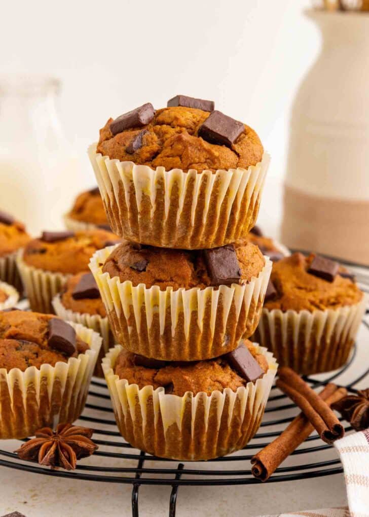 Three pumpkin chocolate chunk muffins stacked on a cooling rack, with more muffins in the background. Cinnamon sticks and star anise are nearby, suggesting a spiced flavor. The scene is bright and inviting.