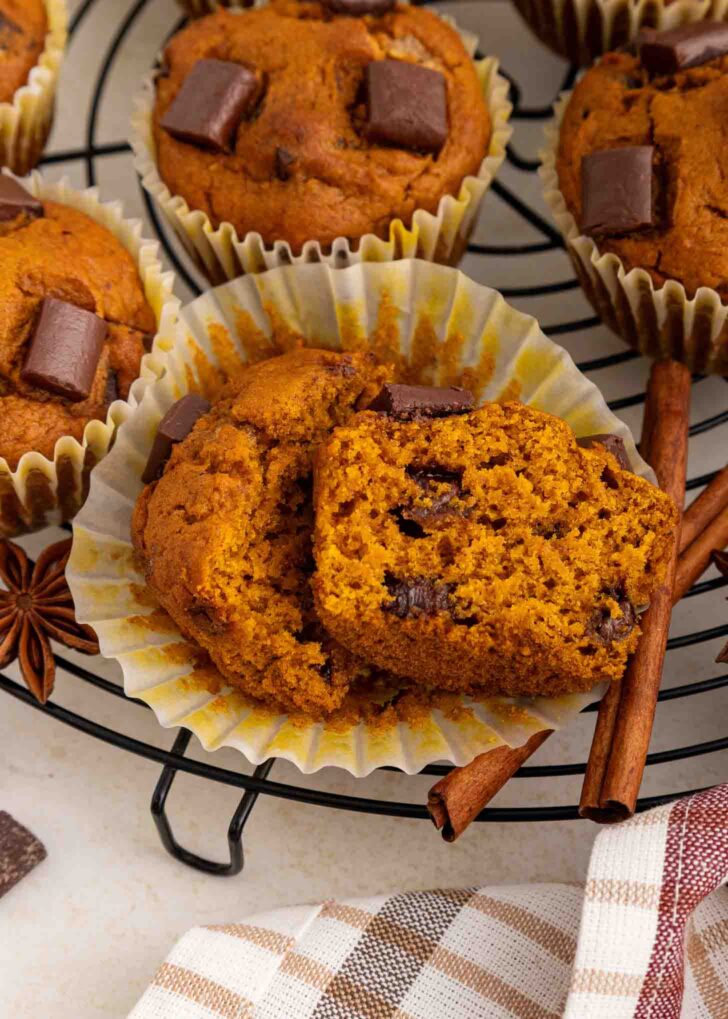 A pumpkin muffin with chocolate chunks is cut in half, resting on a paper liner. Other whole muffins and cinnamon sticks are nearby on a cooling rack.