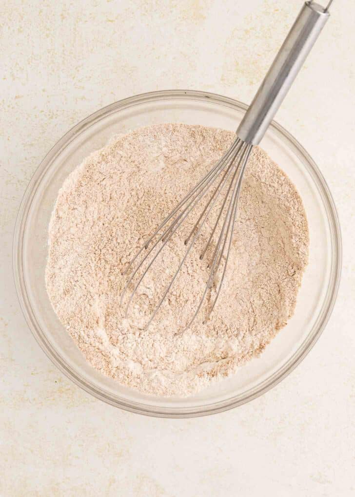 A glass bowl filled with flour mixture sits on a light surface, with a metal whisk resting inside the bowl.