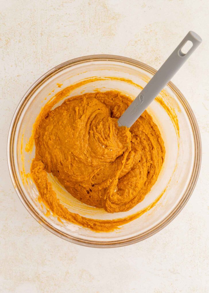 A glass bowl filled with orange pumpkin batter is being mixed with a gray spatula, shown on a light-colored surface.