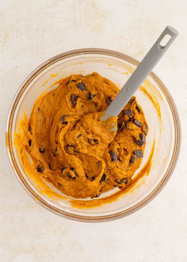 A glass bowl filled with pumpkin-colored cookie dough mixed with chocolate chunks. A gray spatula is resting inside the bowl. The bowl sits on a light-colored surface.