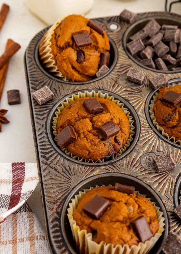 Pumpkin muffins with chocolate chunks in a metal muffin tin, some muffins partially visible, with extra chocolate pieces nearby and a striped cloth in the corner.