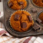A muffin tin with pumpkin muffins topped with chocolate chunks, one muffin has a bite taken out. A checkered cloth is partially visible underneath the tin.