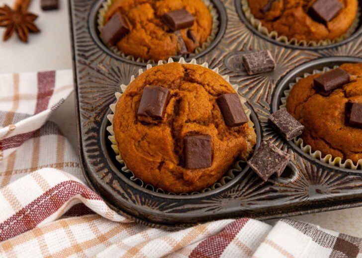 A muffin tin with pumpkin muffins topped with chocolate chunks, one muffin has a bite taken out. A checkered cloth is partially visible underneath the tin.