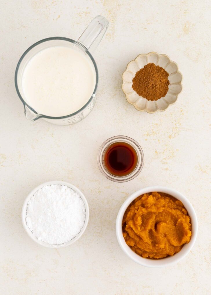 Five recipe ingredients on a white surface: a measuring cup of milk, a small dish of brown spice, a bowl of orange pumpkin puree, a bowl of white powder, and a small glass container with dark liquid.