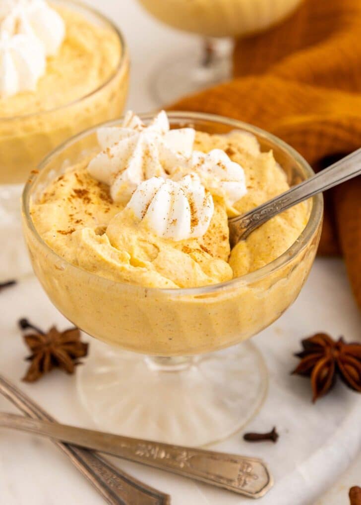 A glass bowl filled with creamy pumpkin mousse, topped with whipped cream and a sprinkle of cinnamon, with a spoon inside. Star anise and cloves are nearby on the table.