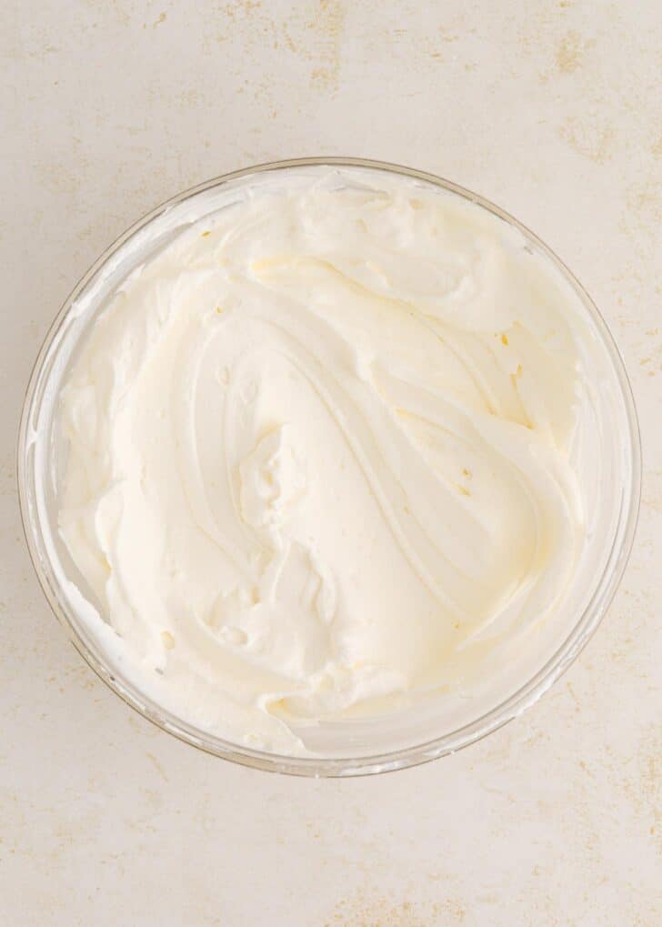 A glass bowl filled with smooth, fluffy whipped cream sits on a light, textured surface.