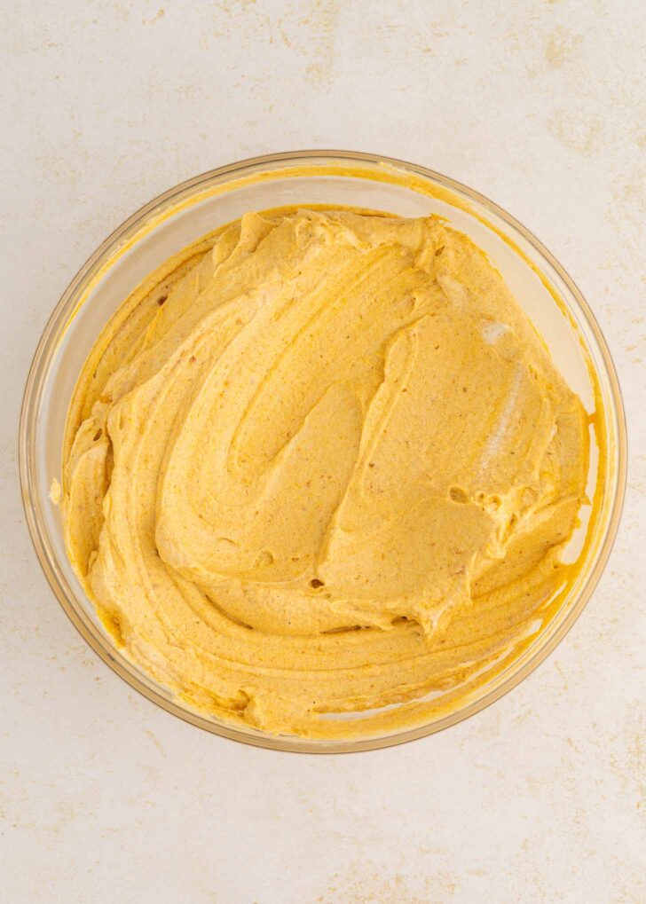 A glass bowl filled with creamy, light orange pumpkin mousse mixture, swirled smoothly on a light-colored surface.