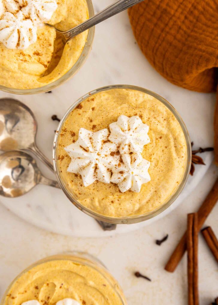 A glass of creamy pumpkin mousse topped with whipped cream and a sprinkle of cinnamon, with a spoon, cinnamon sticks, and cloves nearby on a marble surface.