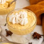 A glass dish filled with creamy, orange pumpkin mousse topped with whipped cream and sprinkled with cinnamon, surrounded by star anise, cloves, spoons, and an orange napkin.