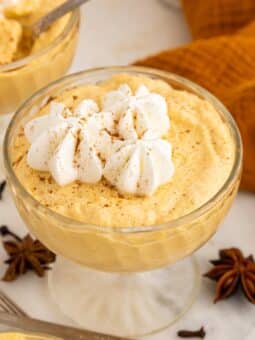 A glass dish filled with creamy, orange pumpkin mousse topped with whipped cream and sprinkled with cinnamon, surrounded by star anise, cloves, spoons, and an orange napkin.