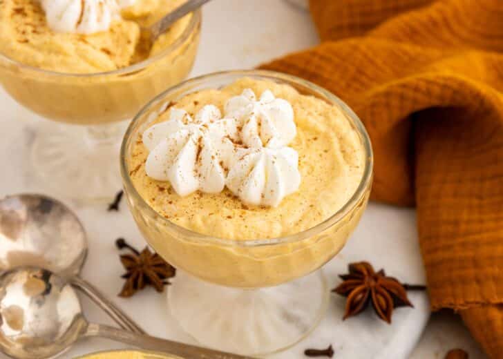 A glass dish filled with creamy, orange pumpkin mousse topped with whipped cream and sprinkled with cinnamon, surrounded by star anise, cloves, spoons, and an orange napkin.
